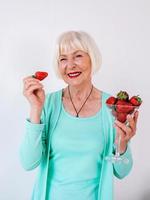 Porträt einer stilvollen, fröhlichen Seniorin in türkisfarbener Kleidung, die Erdbeeren isst. Sommer, Reisen, Anti Age, Freude, Ruhestand, Erdbeeren, Beeren, Vitamine, Freiheitskonzept foto
