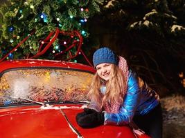 glücklich lächelnde junge kaukasische Frau in Schal, Hut, Jacke, Handschuhe durch den roten Oldtimer und Weihnachtsbaum im Freien. neues Jahr, Spaß, Winterkonzept