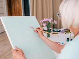 Senior fröhliche Frau mit Brille mit grauem Haar, die mit Bleistiftblumen in der Vase zeichnet. Kreativität, Kunst, Hobby, Berufskonzept foto