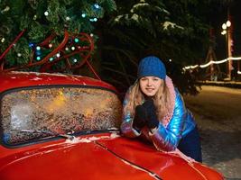 glücklich lächelnde junge kaukasische Frau in Schal, Hut, Jacke, Handschuhe durch den roten Oldtimer und Weihnachtsbaum im Freien. neues Jahr, Spaß, Winterkonzept