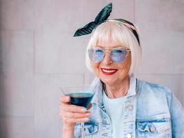 Senior stilvolle Frau mit grauem Haar und in blauer Brille und Jeansjacke mit Glas mit blauem Cocktail. Alkohol, Entspannung, Urlaub, Ruhestandskonzept