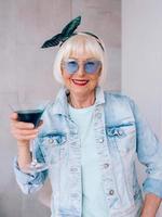 Senior stilvolle Frau mit grauem Haar und in blauer Brille und Jeansjacke mit Glas mit blauem Cocktail. Alkohol, Entspannung, Urlaub, Ruhestandskonzept