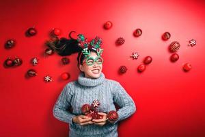 Weihnachten neues Jahr. junge Frau, gekleidet in warmen Pullover mit Requisiten Ball rot mit Weihnachtsschmuck im Urlaub auf glänzendem rotem Hintergrund. Konzept Frohe Weihnachten. foto