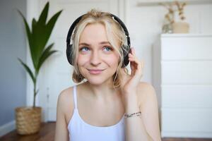 Porträt von gut aussehend jung blond Frau im kabellos Kopfhörer, hört zu zu Podcast oder Lieblings Lied, genießt Musik- im Neu Kopfhörer foto