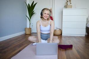 Porträt von Frau während trainieren, Sitzung auf Yoga Matte mit Widerstand Band, Hören zu Anleitung auf Laptop, tragen kabellos Kopfhörer, wiederholen Übungen foto
