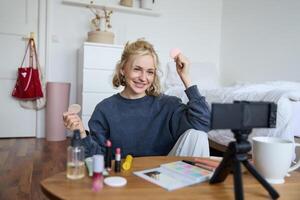 Porträt von jung Frau, Schönheit Inhalt Schöpfer, Sitzung im ein Zimmer im Vorderseite von Digital Kamera, Aufzeichnung bilden Lernprogramm vlog, zeigen kosmetisch Gesichts- Produkte foto