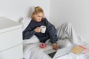 Porträt von jung Frau, Schüler studieren im ihr Bett, entspannend während vorbereiten Hausaufgaben, Essen Krapfen, mit Laptop im Schlafzimmer und Trinken Tee foto