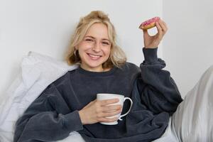 schließen oben Porträt von schön lächelnd, blond Frau, Lügen im Bett, Getränke Tee und isst Krapfen auf ein faul Wochenende oder freier Tag foto