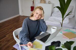 Porträt von lächelnd, schön blond Frau, Schreiben Nieder Anmerkungen, tun Hausaufgaben, studieren von heim, tun Entfernung Lernen, online Kurs, Arbeiten entfernt im ihr Zimmer foto