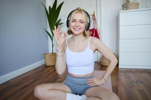 Porträt von süß lächelnd blond Frau tragen kabellos Kopfhörer, trainieren beim Zuhause auf Gummi Yoga Matte, Sitzung im ein Zimmer, zeigen Okay, Ja Hand Zeichen foto