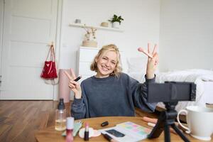 Porträt von jung blond Frau, Teenager Mädchen Aufzeichnungen zum ihr Sozial Medien Konto, zeigt an bilden auf Kamera, empfiehlt Lippenstift zu online Anhänger, schafft Inhalt im ihr Zimmer foto