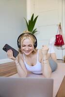 Vertikale Schuss von jung Fitness Frau im Kopfhörer, Lügen auf Gummi Matte, entspannend nach trainieren Ausbildung Sitzung, halten Smartphone, lächelnd beim Kamera foto