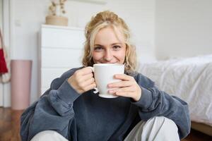 Bild von jung Teenager Mädchen Sitzung im ihr Schlafzimmer auf Boden, Trinken Tasse von Tee und genießen Tag beim heim, lächelnd und suchen beim Kamera foto