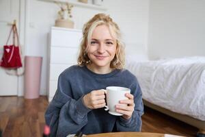 Bild von jung Teenager Mädchen Sitzung im ihr Schlafzimmer auf Boden, Trinken Tasse von Tee und genießen Tag beim heim, lächelnd und suchen beim Kamera foto