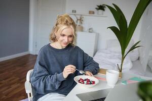 Porträt von jung offen Mädchen Aufpassen s auf Laptop, genießen Film und Essen im Vorderseite von Laptop, haben Frühstück und starren beim Bildschirm foto