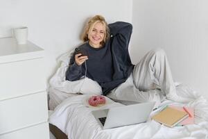 Porträt von jung Frau, Schüler studieren im ihr Bett, entspannend während vorbereiten Hausaufgaben, Essen Krapfen, mit Laptop im Schlafzimmer und Trinken Tee foto