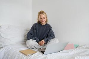Porträt von jung weiblich Student, Frau studieren online, E-Learning auf ihr Laptop, Sitzung auf Bett mit Notizbuch und suchen beim Bildschirm, chatten, verbindet zu ein im eigenen Tempo Kurs foto
