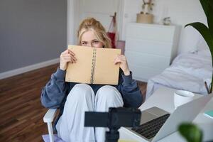 Porträt von süß blond Frau, sitzt im Vorderseite von Digital Kamera und Laptop im ihr Zimmer, Abdeckungen Gesicht mit Notizbuch Tagebuch, Aufzeichnungen von Sie selber zum Lebensstil vlog foto