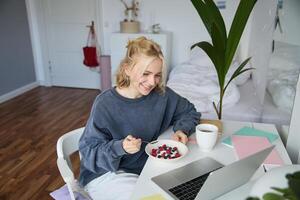 Bild von lächelnd jung Frau Aufpassen s auf Laptop während Essen Frühstück und Trinken Tee, suchen beim Bildschirm, Sitzung im ihr Zimmer foto