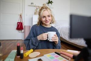 Porträt von süß blond Frau, Inhalt Hersteller, vlogger Aufzeichnung auf Digital Kamera im Schlafzimmer, Trinken Tasse von Tee und reden zu Publikum foto