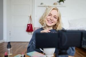 schließen oben Porträt von jung Mädchen Vlogger, Teenager Aufzeichnungen Blog Über bilden, Verwendet Kamera auf Stabilisator, Sitzung auf Fußboden in der Nähe von Kaffee Tisch, bewirbt sich Wimperntusche und kosmetisch Produkte foto