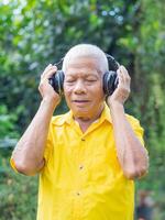 Senior Mann tragen kabellos Kopfhörer zum Hören zu ein Liebling Lied, und Augen geschlossen während Stehen im ein Garten. Konzept von alt Menschen und Entspannung foto