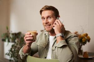 Bild von lächelnd, gut aussehend jung blond Mann mit Telefon, Trinken Kaffee im Cafe, reden zu jemand Über das Telefon, antworten ein Anruf foto