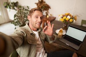 Porträt von jung gut aussehend Mann, Cafe Besucher nehmen Selfie auf Smartphone Anwendung, zeigen Okay, gut Empfehlung Zeichen, Blogger Preise Kaffee Geschäft, nickt im die Genehmigung foto