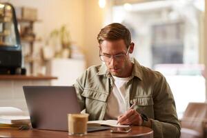 Bild von konzentriert jung Mann im Gläser, Sitzung im Cafe, Herstellung Anmerkungen, studieren, Teilnahme online Kurs, Lernen auf Fernbedienung von ziemlich Kaffee Geschäft foto