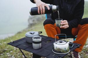 gießen Tee von ein Thermosflasche in ein Becher im Natur, ein Tourist gießt ein heiß trinken in ein Glas, ein Berg Wanderung Frühstück im das Nebel, Tourist Utensilien Camping auf ein Felsen foto