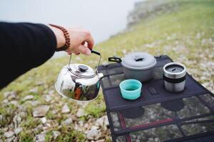 Frühstück von ein Tourist auf ein Wanderung Sitzung auf das Berg beim das Tisch, Hand braut Tee Sieden Wasser im ein Becher, ein Metall Wasserkocher, nehmen ein Hand durch das handhaben, zwei Gläser, ein schwenken zum Essen foto