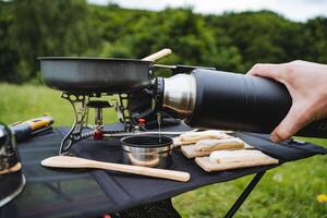 ein Person hält im seine Hand ein Thermosflasche mit Tee, gießen Wasser in ein Becher im Natur, Essen auf ein Wanderung, ein Camping Tisch, Frühstück im Natur, Tourist Teller, kompakt Ausrüstung. foto