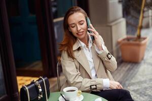 lächelnd modisch Frau Sitzung beim Cafe Terrasse reden auf Telefon während Trinken Kaffee foto
