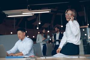 lächelnd reifen Geschäft Frau Sitzung auf Tabelle und reden Telefon während Treffen im modern Büro foto