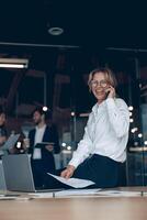 lächelnd reifen Geschäft Frau Sitzung auf Tabelle und reden auf Telefon im modern Büro foto