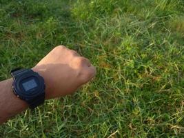 ein Mann, der seine Uhr mit geballten Fäusten auf einem Grasgartenhintergrund zeigt foto