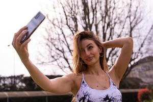 jung charmant blond sportlich Frau im Sportbekleidung posieren nehmen ein Selfie draußen foto