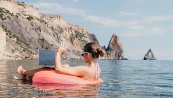 Frau Freiberufler funktioniert auf Laptop Schwimmen im Meer auf Rosa aufblasbar Ring. glücklich Tourist im Sonnenbrille schwebend auf aufblasbar Krapfen und Arbeiten auf Laptop Computer im Ruhe Ozean. Fernbedienung Arbeiten irgendwo foto