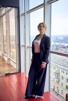 attraktiv Mitte Alter Frau im Jacke und Weiß T-Shirt posieren im modern Büro mit Panorama- Fenster foto