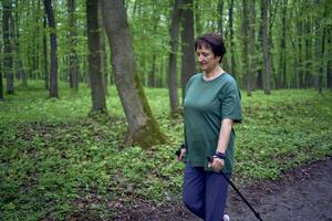 Alten Frau ist beschäftigt, verlobt im nordisch Gehen mit Stöcke im das Frühling Wald foto