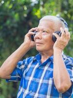 Senior Mann tragen kabellos Kopfhörer Hören zu ein Liebling Lied und suchen oben während Stehen im ein Garten. Raum zum Text. Konzept von alt Menschen und Entspannung foto