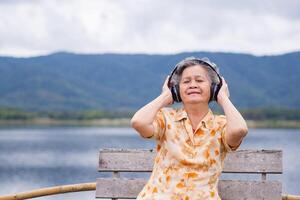 Porträt von ein glücklich Senior Frau tragen kabellos Kopfhörer Hören zu Musik- zum entspannend während Sitzung Seite das See. Konzept von alt Menschen und Entspannung foto