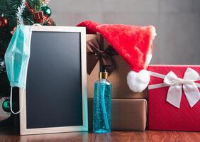 klein Tafel, Maske, Alkohol Gel, und Geschenk Kisten auf hölzern Tabelle mit ein Weihnachten Baum und Jahrgang Mauer Hintergrund. Raum zum Text. Konzept von Weihnachten, Neu Jahr Festival und Gesundheitswesen foto