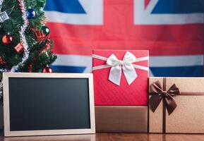 klein Tafel und Geschenk Kisten auf hölzern Tabelle mit ein Weihnachten Baum und vereinigt Königreich Flagge im das Hintergrund. Raum zum Text. Konzept von Weihnachten und Neu Jahr Festival foto