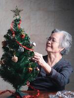 Porträt von ein Alten asiatisch Frau dekorieren das Weihnachten Baum während Sitzung im ein Leben Zimmer beim heim. Raum zum Text. Konzept von alt Menschen und Weihnachten und glücklich Neu Jahr Festival foto