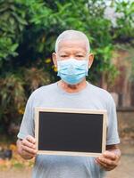 Senior Mann tragen ein Gesicht Maske und halten ein leer Tafel während Stehen im ein Garten. Maske zum schützen Virus, Coronavirus, Pollen Körner, und mehr. Konzept von alt Menschen und Gesundheitswesen foto