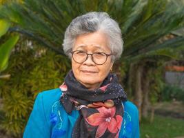 Porträt von ein Alten asiatisch Frau tragen Gläser, lächelnd, und suchen beim das Kamera während Stehen im ein Garten. Konzept von alt Menschen und Gesundheitswesen foto