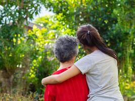 zurück Aussicht von ein jung Frau umarmen ihr Oma im das Garten. Konzept von alt Menschen und Gesundheitswesen foto