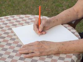 Hände von ein Senior Frau Schreiben ein Buch während Sitzung auf ein Stuhl im das Garten. Nahansicht Foto mit Kopieren Raum zum Text. Konzept von alt Menschen und Gesundheitswesen