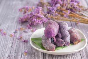 ein Stapel von reif lila Süßkartoffeln ist platziert auf ein Teller auf ein hölzern Tabelle mit verschwommen lila Blumen im das Hintergrund. Raum zum Text. Konzept von Gesundheit Obst foto